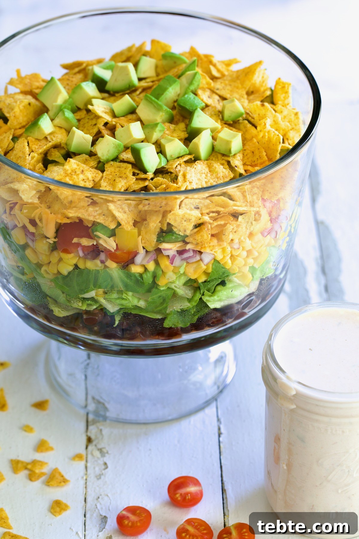 Layered Southwestern Salad with Corn, Black Beans, and Avocado 2 Southwestern Layered Salad in glass footed bowl with salad dressing in small glass jar next to it.