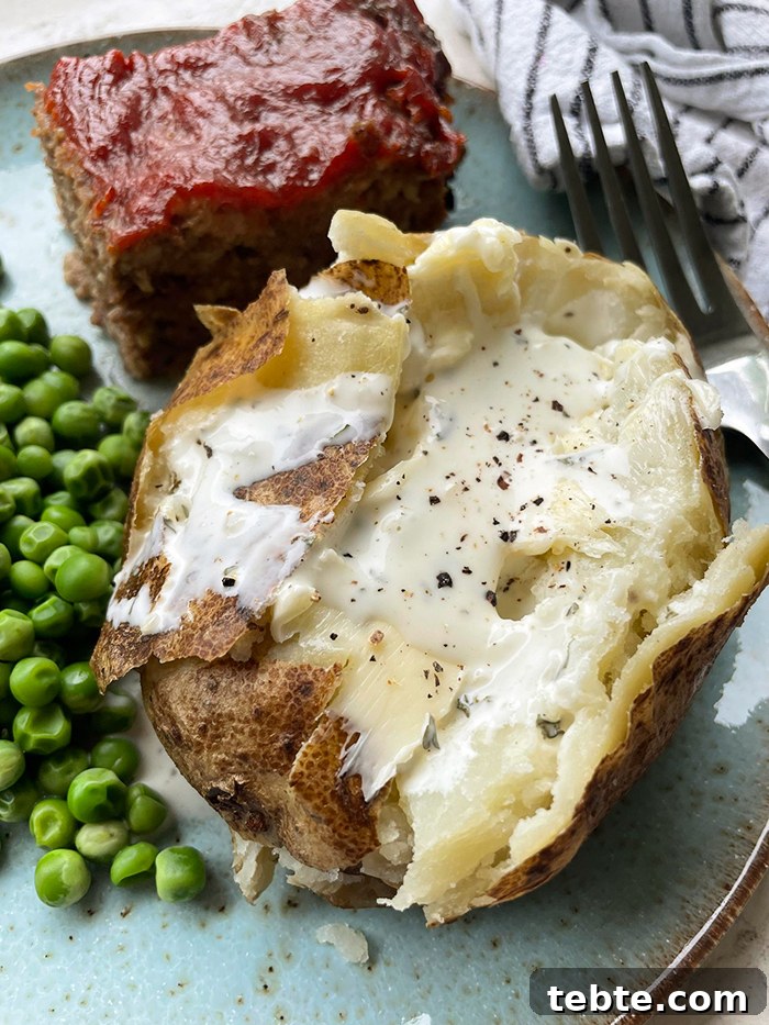 Fancy-Looking Christmas Eve Dinner Ideas for Busy Families 14 A large baked potato cut open with butter, salt and pepper on a dinner plate with a slice of meatloaf and a serving of green peas. A fork head is on the plate on the right of the potato.