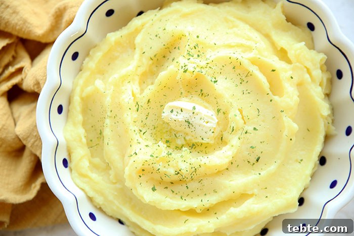 Fancy-Looking Christmas Eve Dinner Ideas for Busy Families 16 Mashed potatoes with a small scoop of butter in the middle with parsley sprinkled on top all in a white Polish potter bowl with scallops, blue trim at the top, and dark blue polkadots throughout the bowl. On the left side of the bowl is a cloth yellow napkin scrunched up.