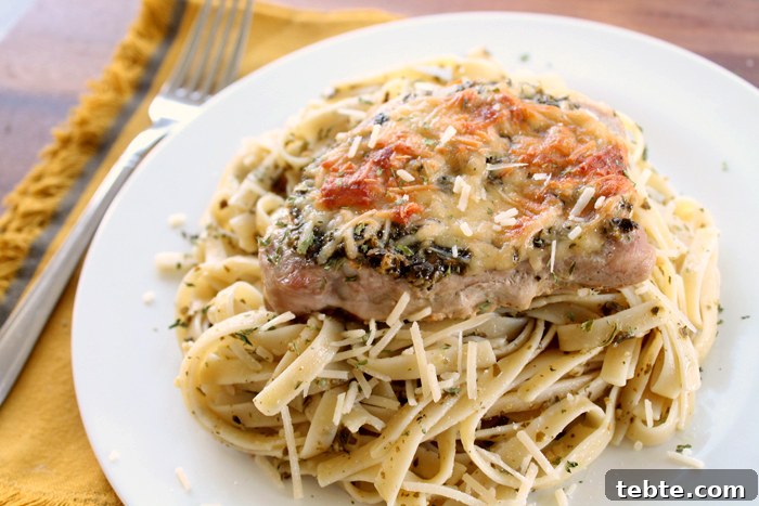 Fancy-Looking Christmas Eve Dinner Ideas for Busy Families 6 Pesto Parmesan Pork chop served on a bed of linguini pasta on a white plate with a fork on the left sitting on a mustard yellow napkin.