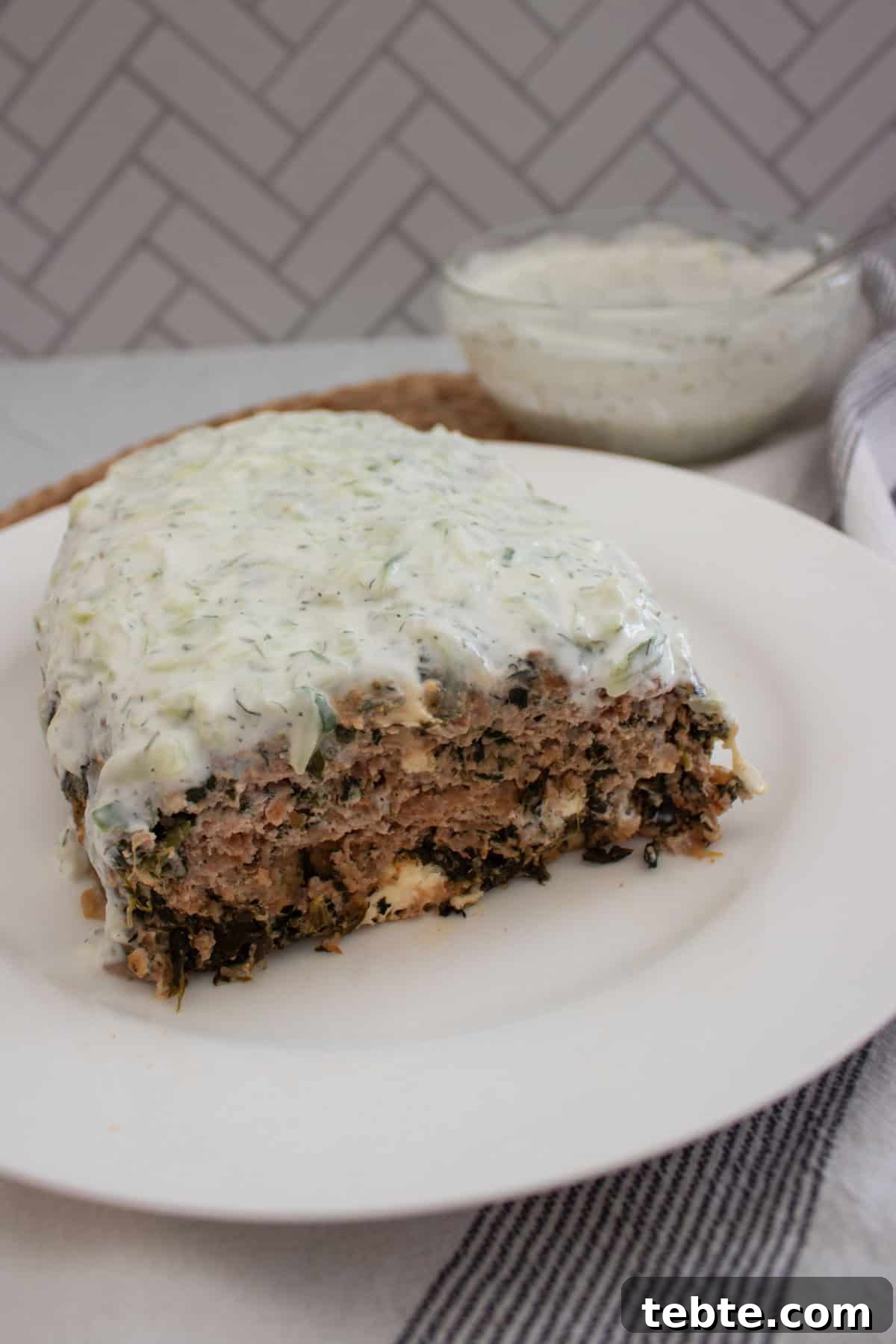 Savory Greek Meatloaf with Spinach and Feta 2 A sliced meatloaf filled with herbs and greens, topped with a creamy white sauce, is served on a white plate. A bowl of extra sauce and a striped napkin are in the background.
