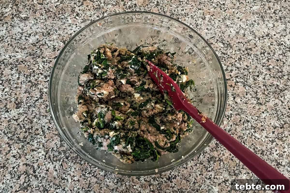 Savory Greek Meatloaf with Spinach and Feta 4 A glass bowl filled with a mixture of cooked greens, browned meat, and white cheese on a speckled countertop. A red mixing spoon rests in the bowl.