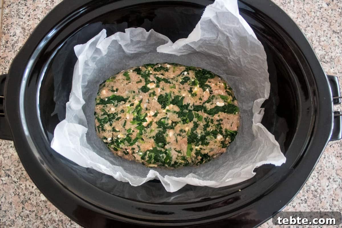 Savory Greek Meatloaf with Spinach and Feta 5 A raw spinach and meat mixture is shaped into a loaf and placed on parchment paper inside a black slow cooker, ready to be cooked. The slow cooker sits on a speckled countertop.