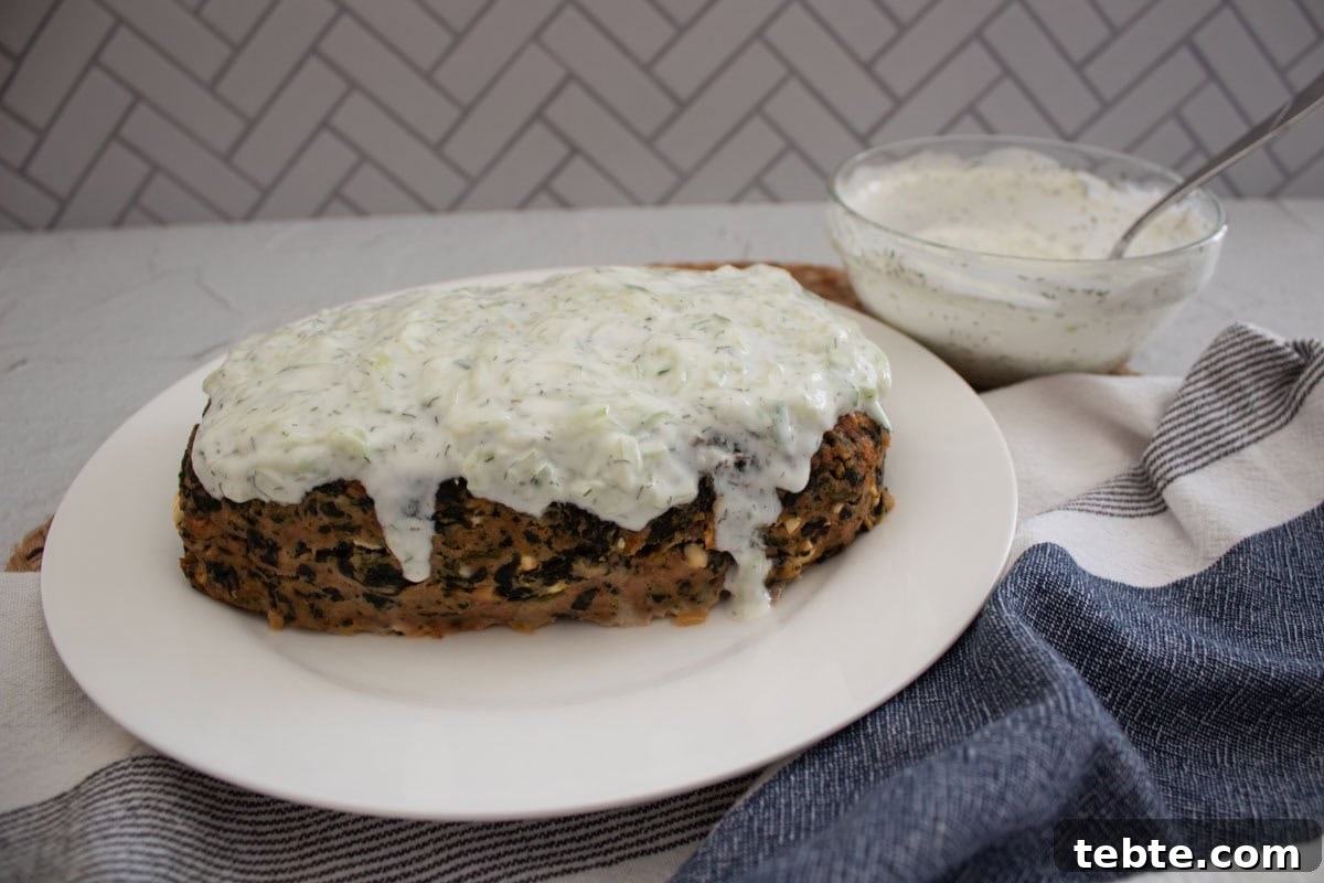 Savory Greek Meatloaf with Spinach and Feta 6 A turkey meatloaf with spinach topped with creamy white sauce on a white plate, with a bowl of extra sauce and a spoon beside it, set on a light table with a blue-striped napkin.