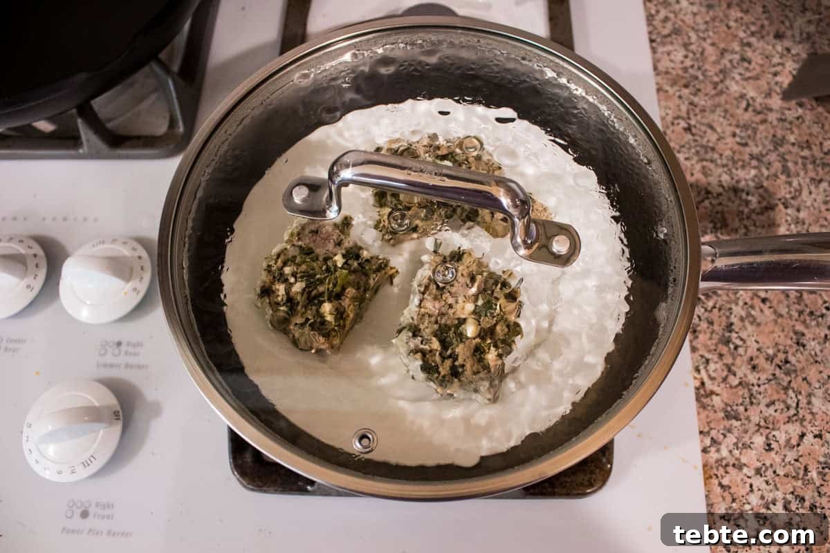 Savory Greek Meatloaf with Spinach and Feta 8 A covered pan on a stove contains three pieces of turkey meatloaf on a heat safe plate. The pan’s transparency reveals the contents, and stove knobs and a speckled countertop are visible nearby.